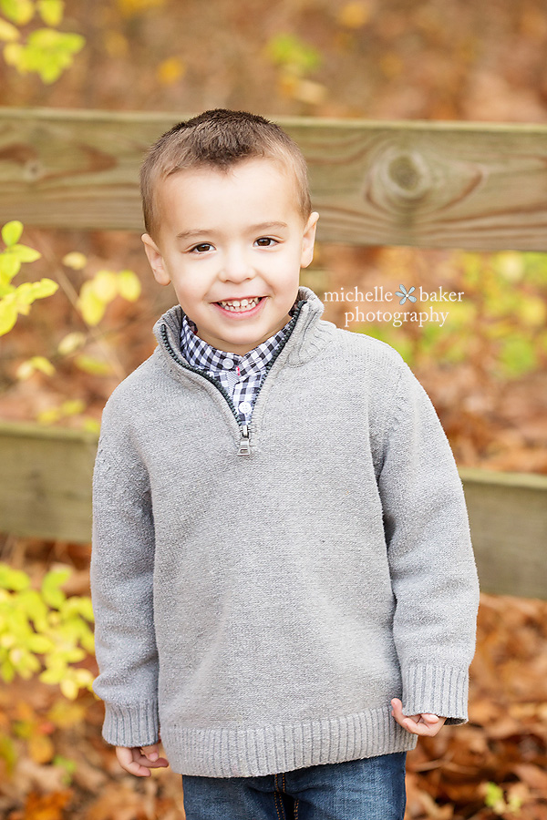 Little boy in grey sweater smiling leaning on a fence